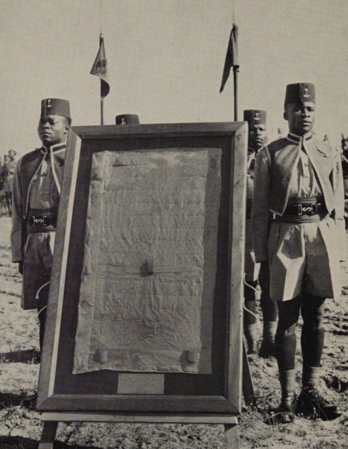 A black-and-white photograph of a framed cloth flag on an easel between two soldiers in fezzes.