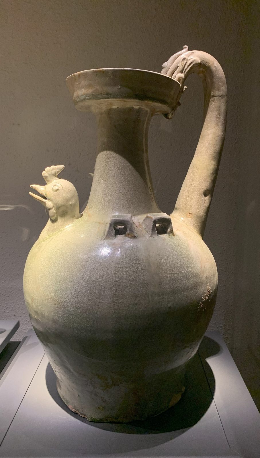 A pale green glazed ewer with a chicken-head spout and a tall curved handle, in a dark museum case.