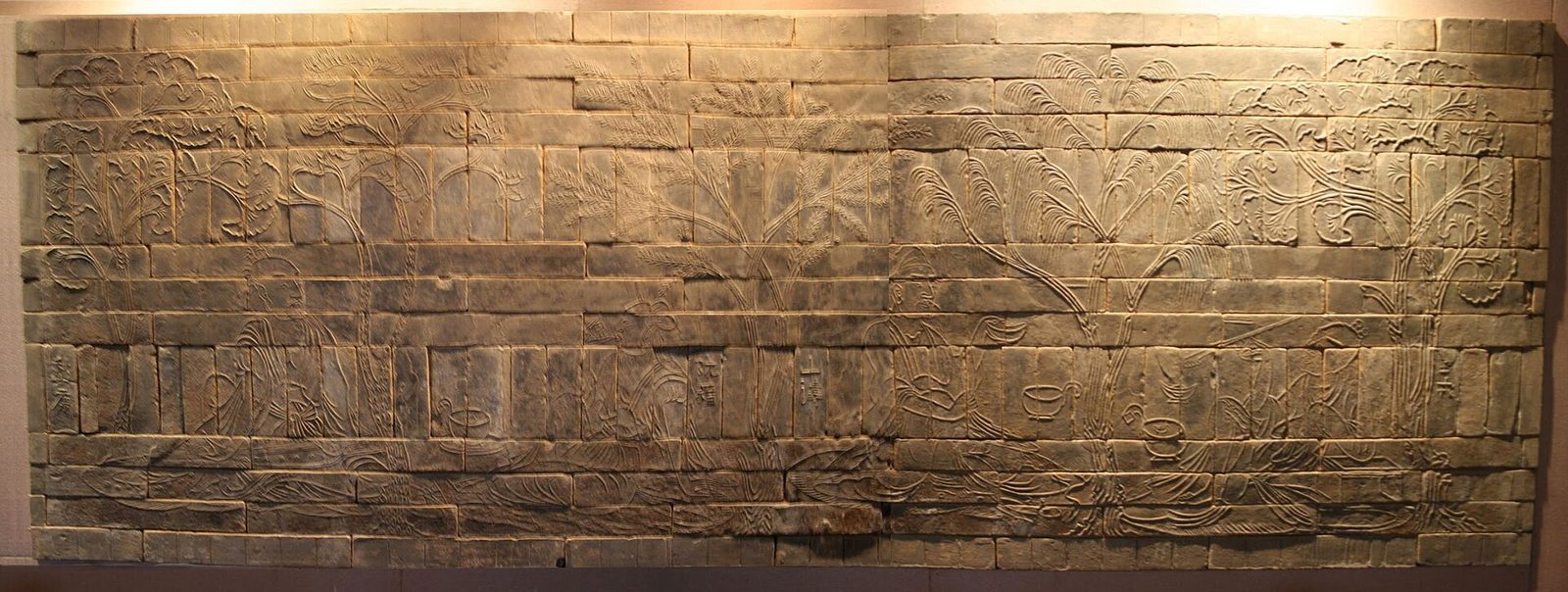 A long wall of moulded bricks forming a relief of seated figures between trees.