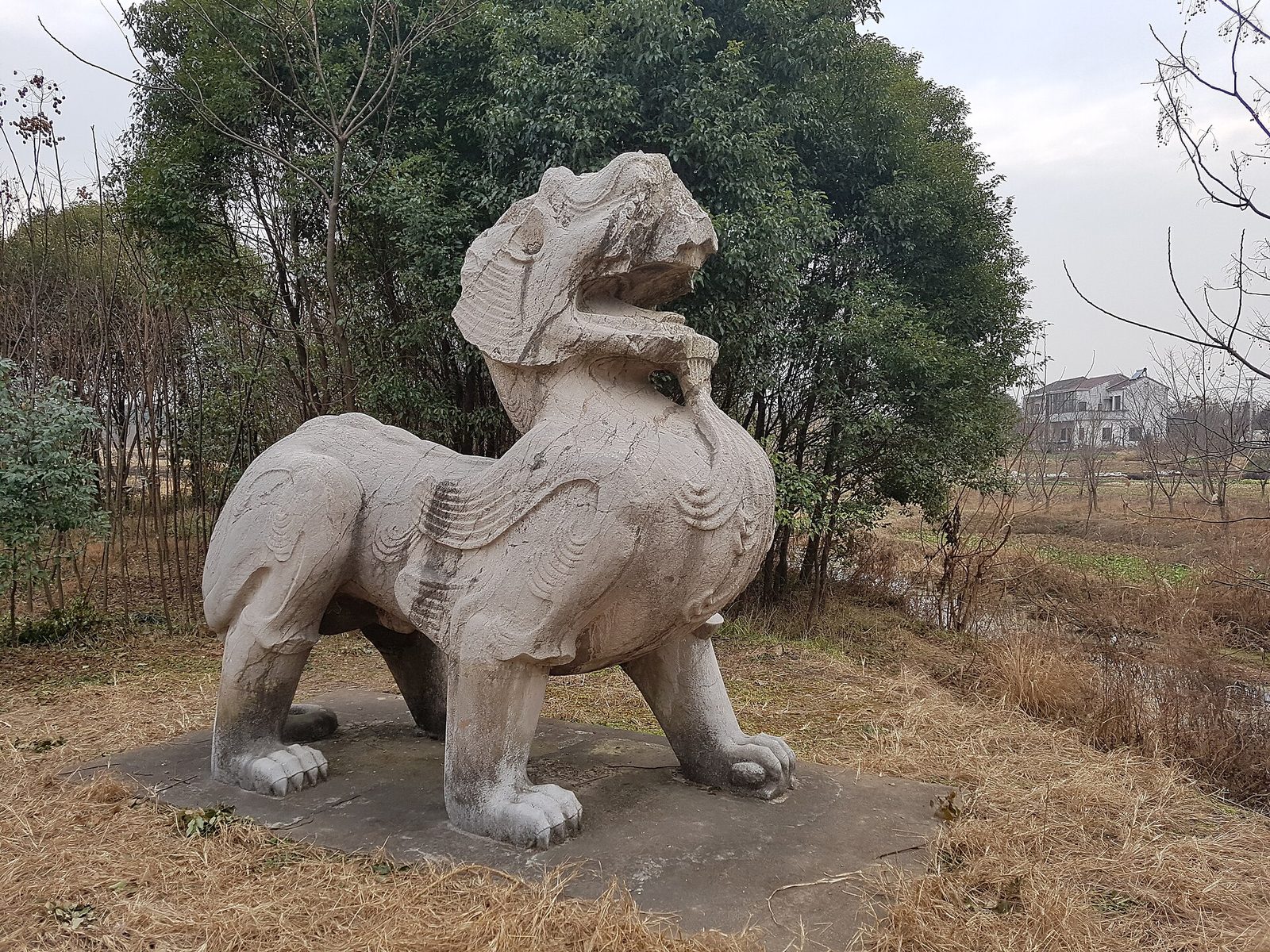 A weathered stone winged beast in mid-stride on a stone base among trees and dry grass.