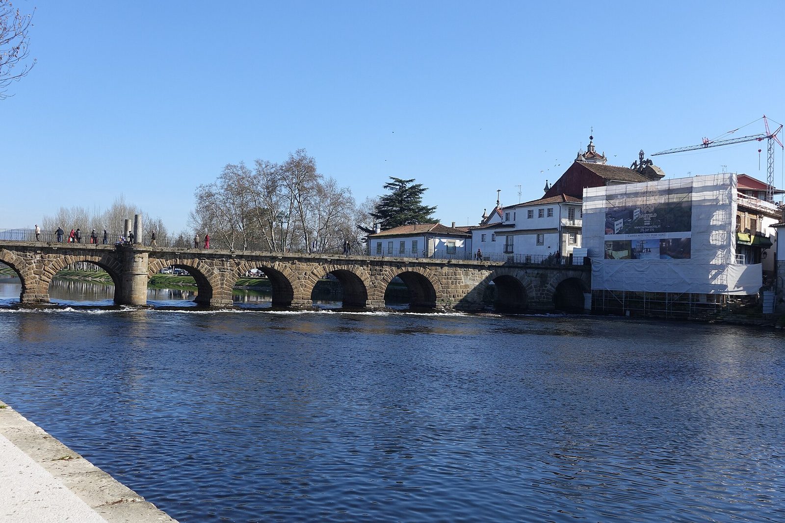 A long low Roman bridge of many shallow arches crossing a wide green river, with a town of white houses and red roofs along the far bank.