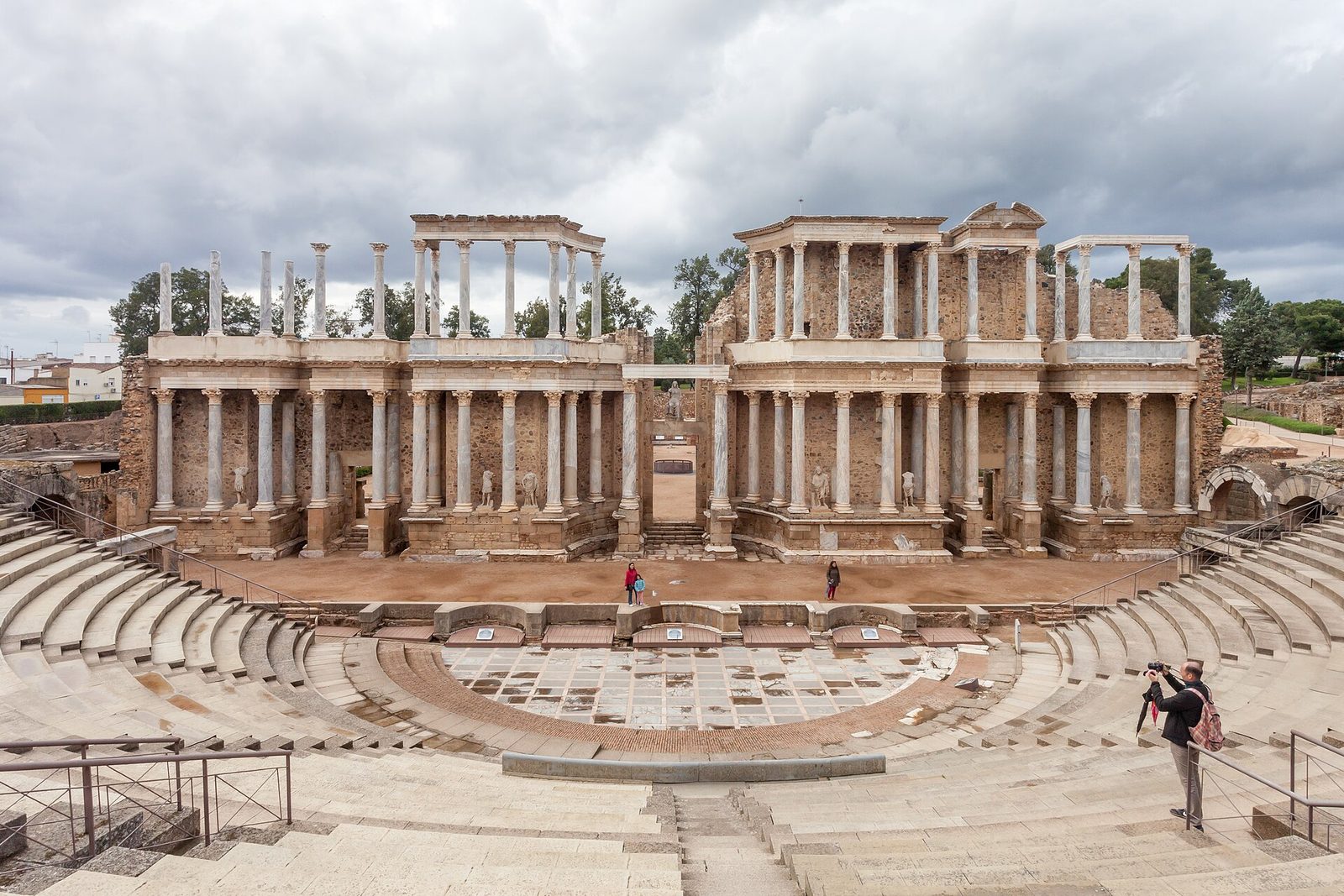A Roman theatre with a tall two-storey stage front of columns and statues standing behind a semicircle of stone seating cut into a slope.