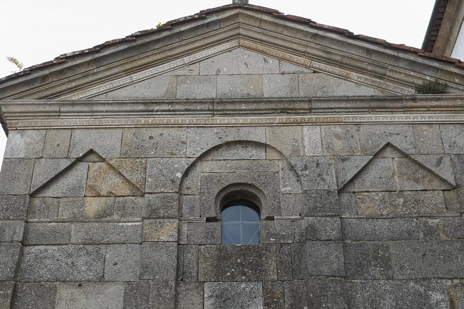 A small cross-shaped stone chapel with low gabled arms and blind arcading round its walls, standing among trees beside a larger later church.