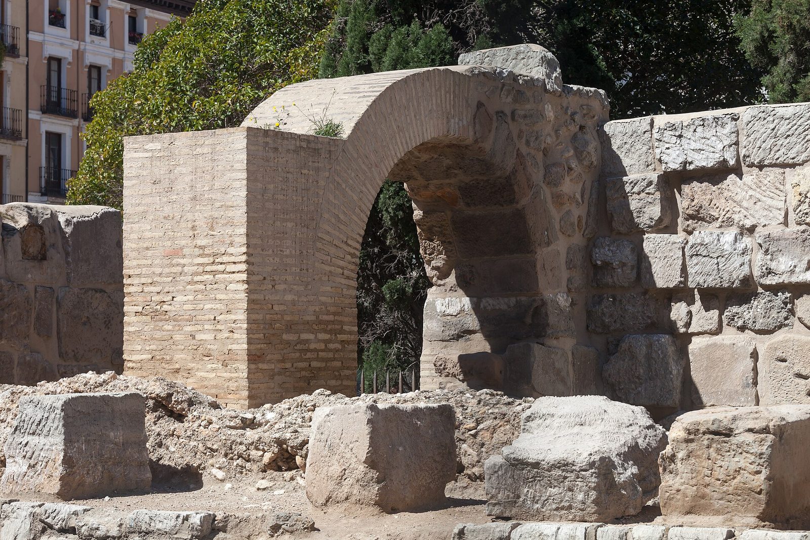 A thick stretch of Roman city wall in large stone blocks with a round tower at one end, standing in a paved square below modern apartment blocks.