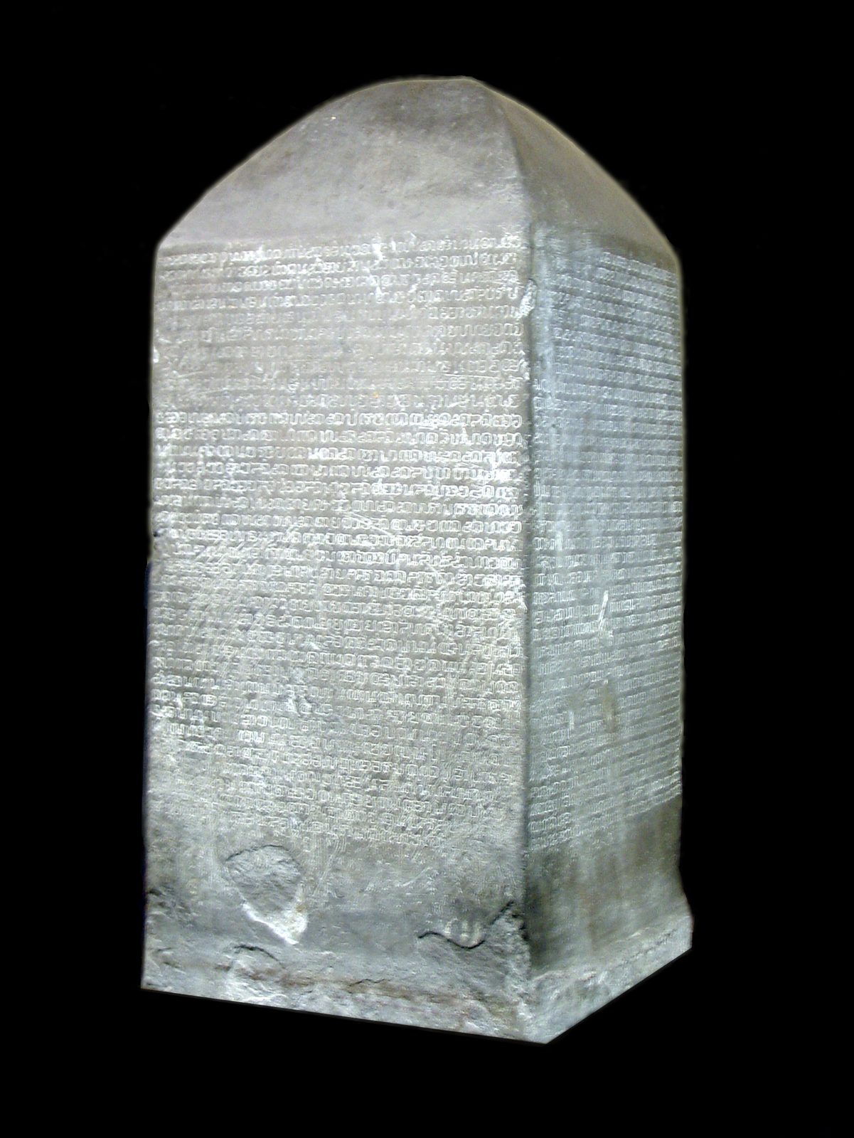 A four-sided stone pillar covered in lines of early Thai script, against black.