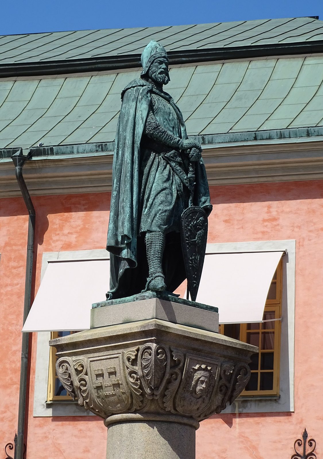 A bronze statue of a man in a long robe and helmet holding a shield, on a carved pedestal before a pink building.