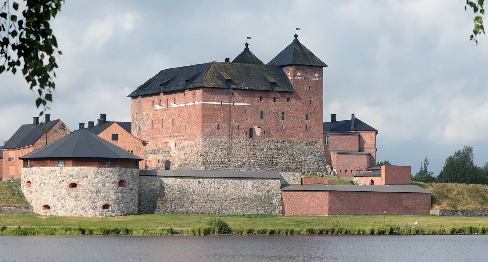 A red brick castle with steep roofs and round towers, seen across water on a summer day.