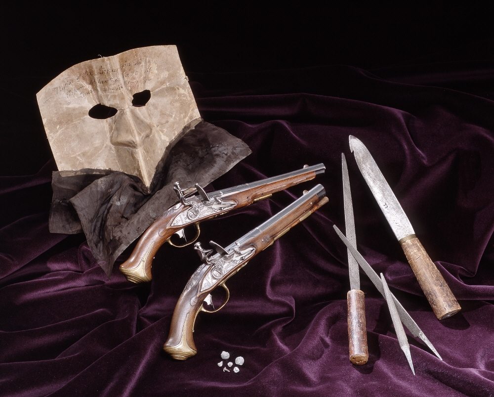 A paper mask, two flintlock pistols and a knife laid out on purple velvet, with small lead shot beside them.
