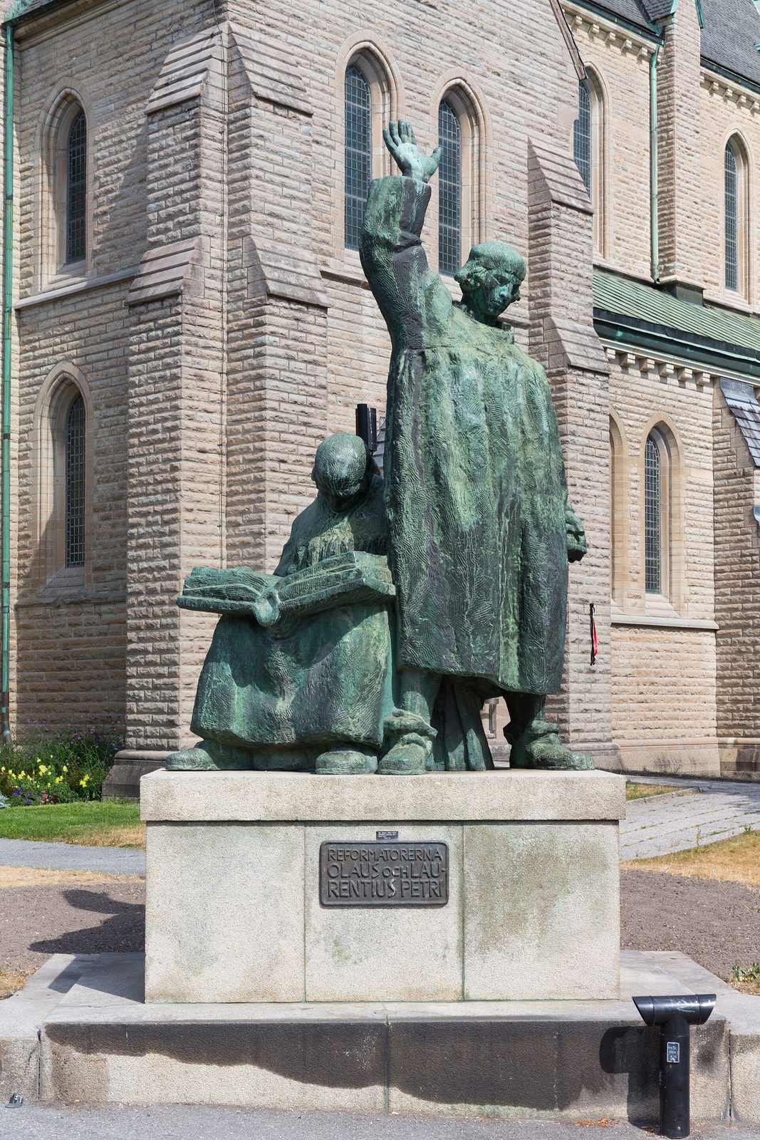 A bronze group of two standing reformers, one with a raised arm, on a stone plinth before a church.