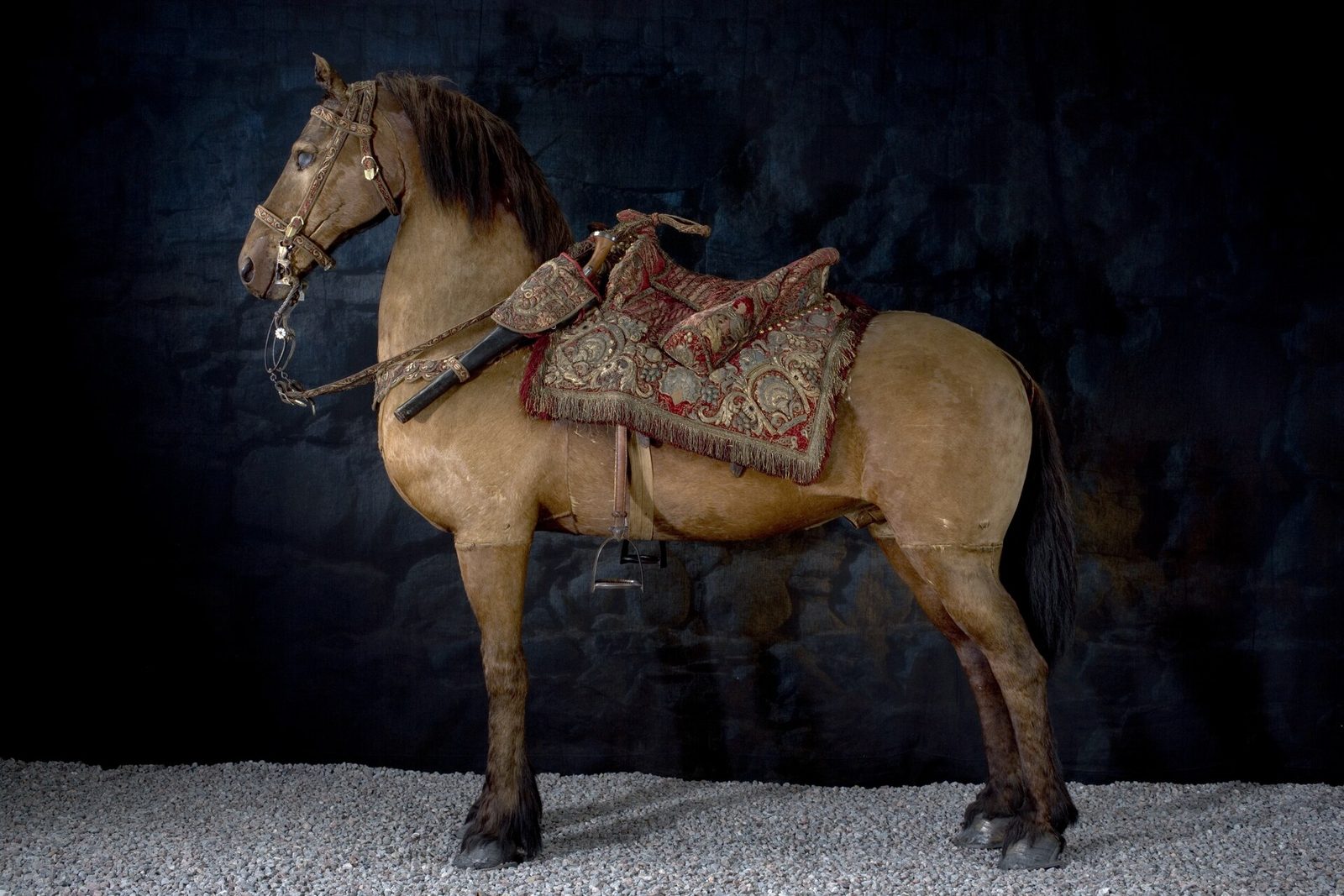 A preserved pale brown horse wearing an embroidered red saddle cloth, standing against a dark background.