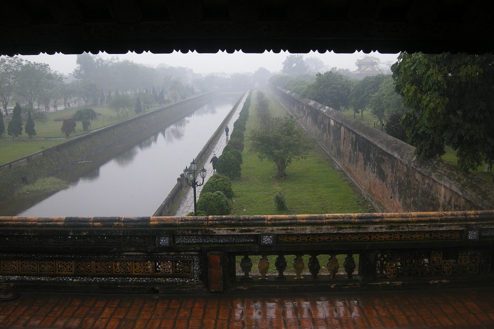 A misty moat between grassy banks and a long brick wall, seen from a covered gallery.