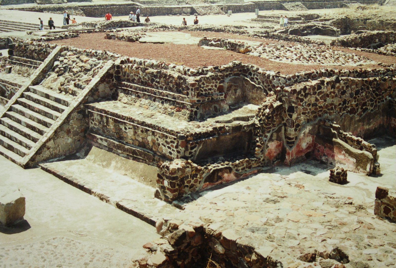 Excavated stone platforms and stairways with traces of red paint beside a broad paved way.