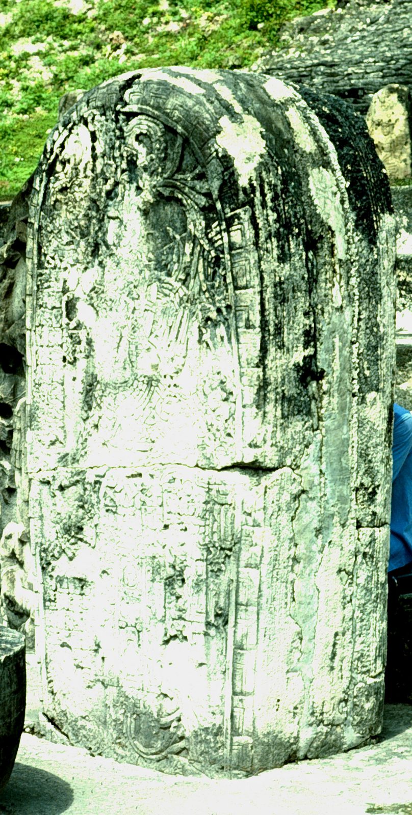 A weathered stone stela with faint carving, streaked with green and white lichen.