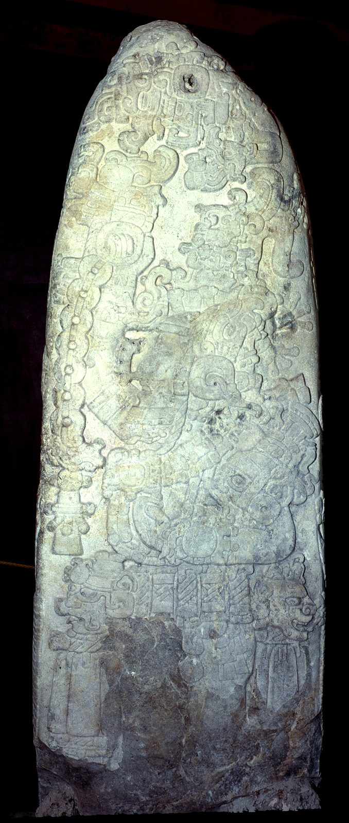 A tall pale stone stela carved all over with a richly dressed figure and glyphs, against black.