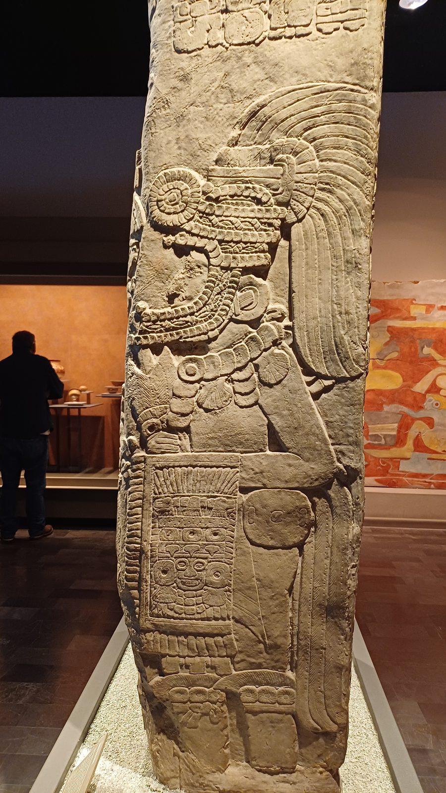 The narrow side of a carved stela showing a standing warrior in a tasselled headdress holding a shield.