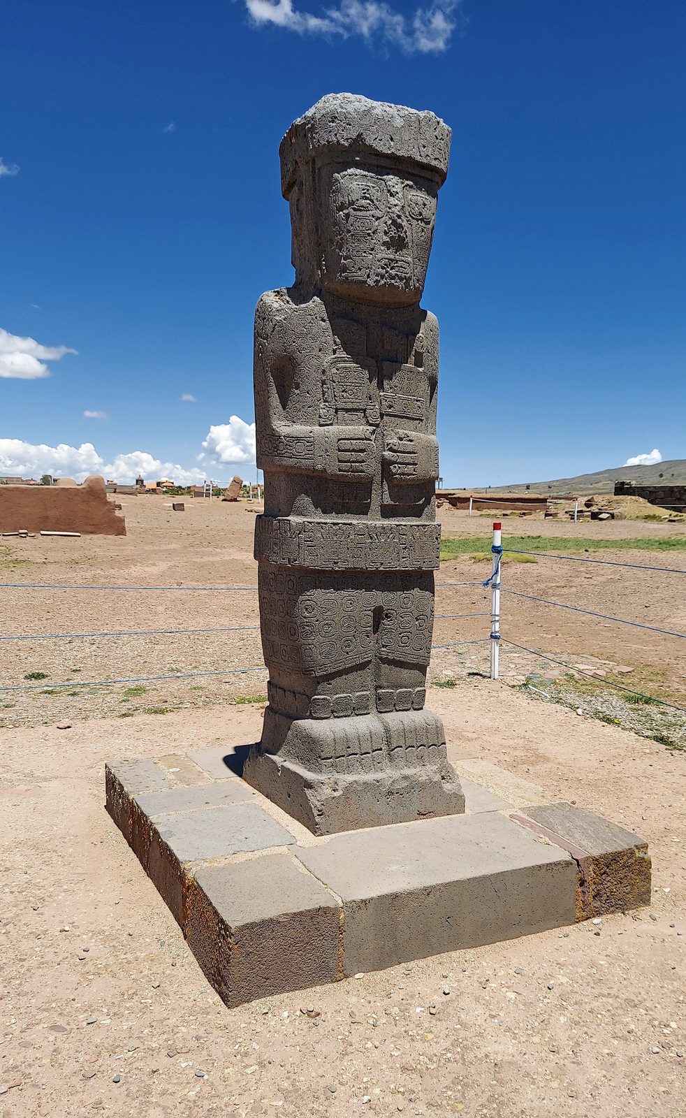 A rectangular stone statue of a standing figure holding objects to its chest, on a stone base under a blue sky.