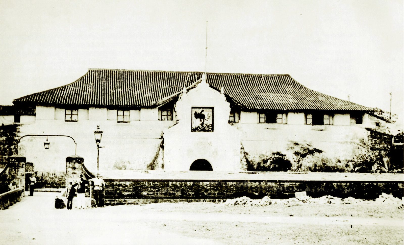 An old photograph of a low fortified gatehouse with a tiled roof and a central arched gate, an open space in front.