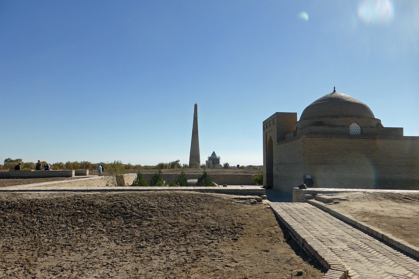 A tall slender brick minaret and a domed mausoleum standing on a flat bare plain.