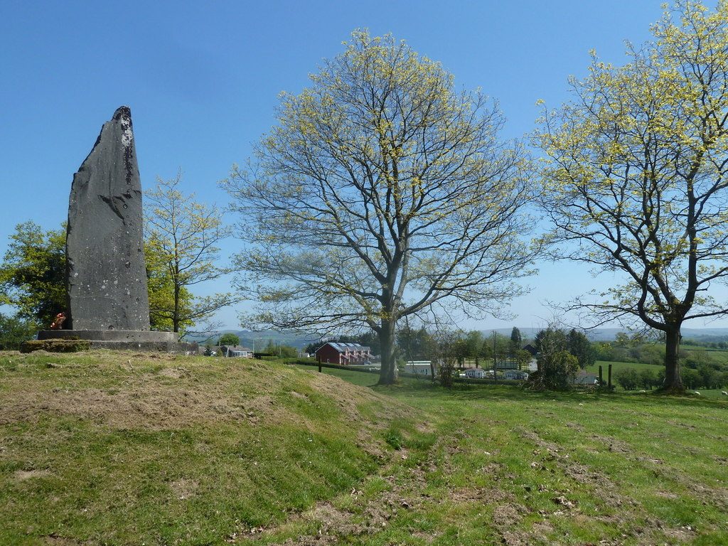 A tall rough stone monolith on a grassy mound among trees.