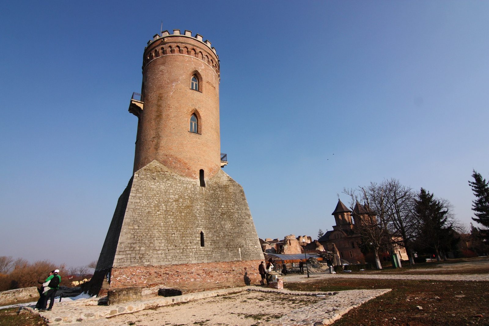 A tall round brick tower on a stone base, beside the ruins of a palace.