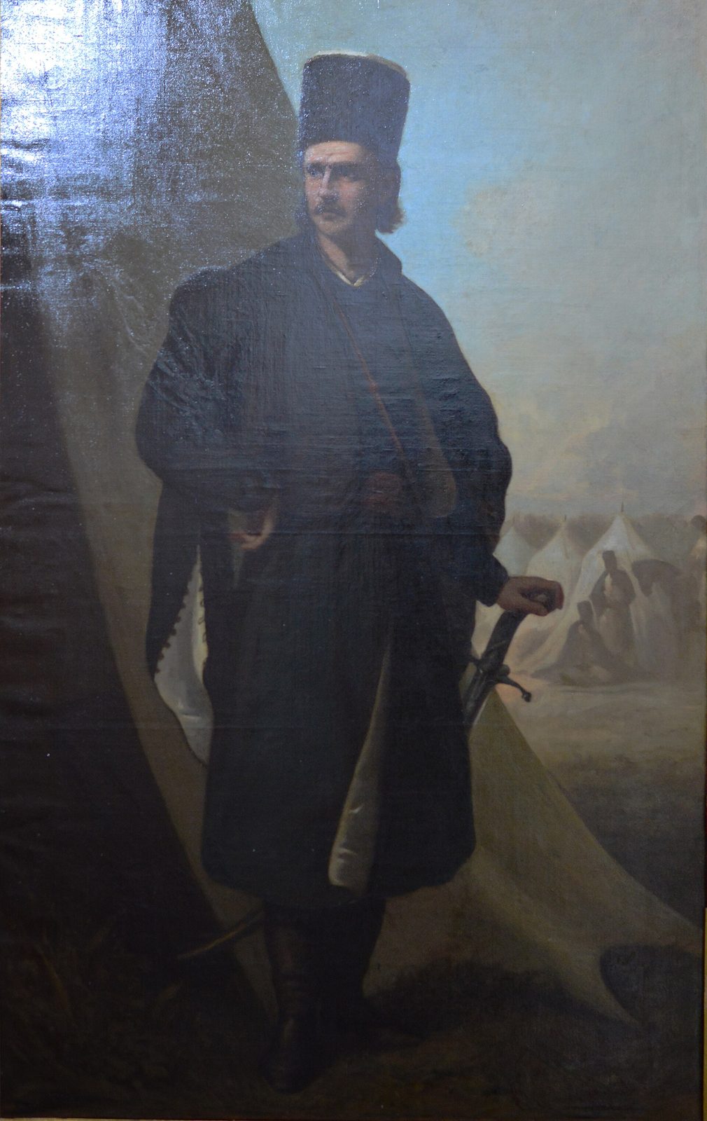 A painting of a bearded man in a dark cloak and tall black cap, standing with a sword before a tent.