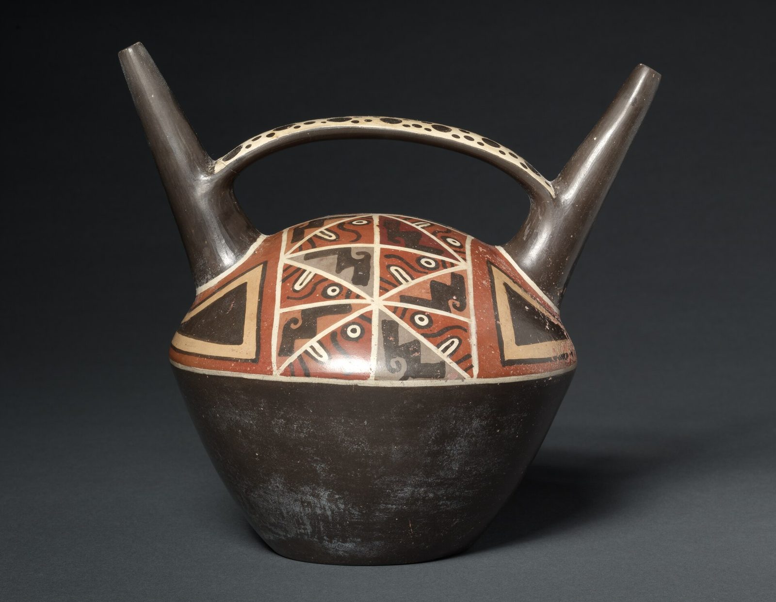 A dark ceramic bottle with two spouts joined by a bridge, its shoulder painted with a band of faces and stepped frets.