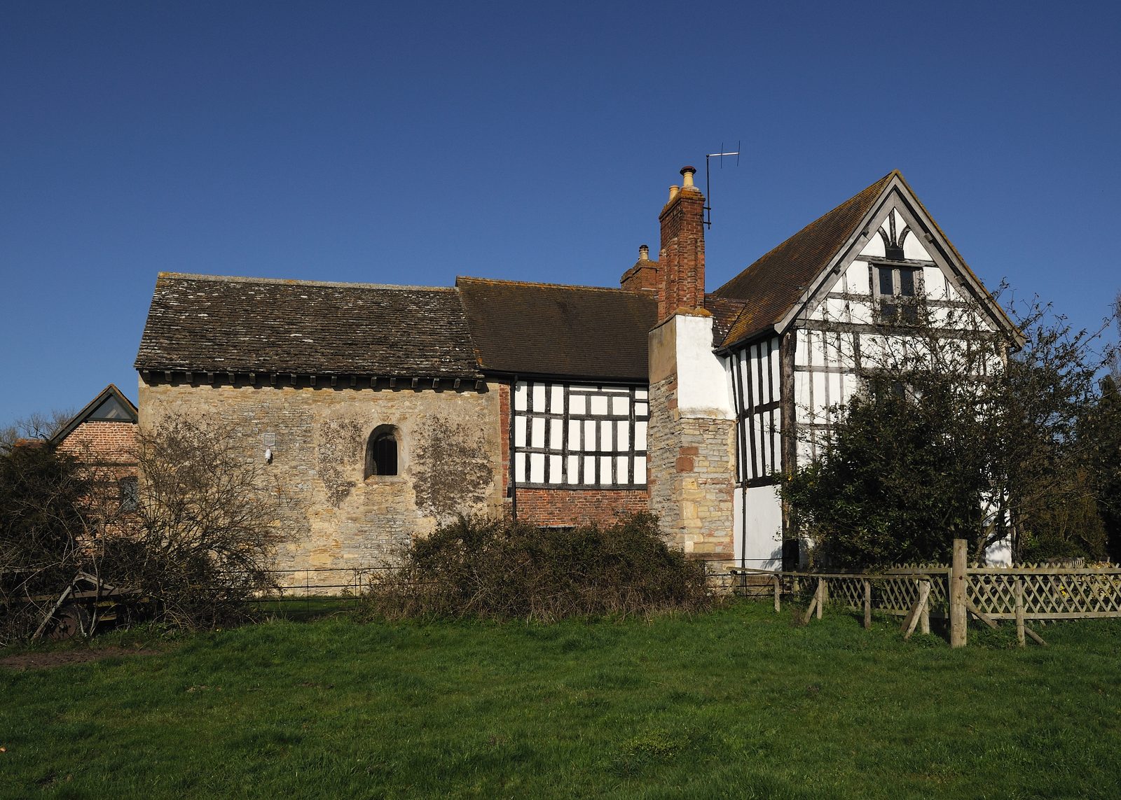 A small stone chapel joined to a timber-framed house, on a lawn.
