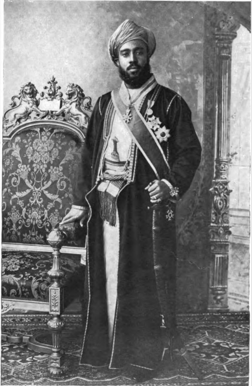 A photograph of a bearded man in a turban and long robe with a sash and orders, standing beside a carved chair.