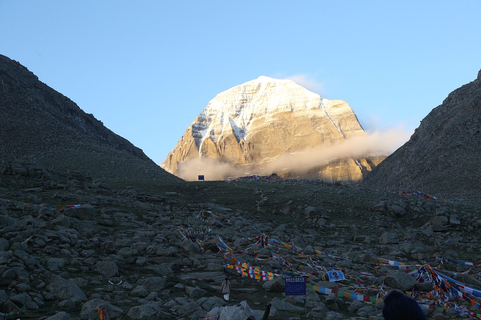 A snow-capped mountain rising at the head of a stony valley, prayer flags strung across the foreground.