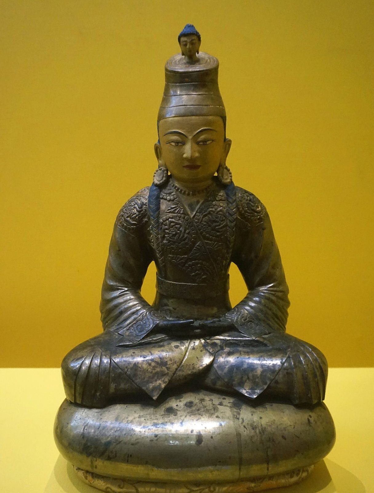 A gilt metal statue of a seated king with a tall turban, hands in his lap, against a yellow wall.