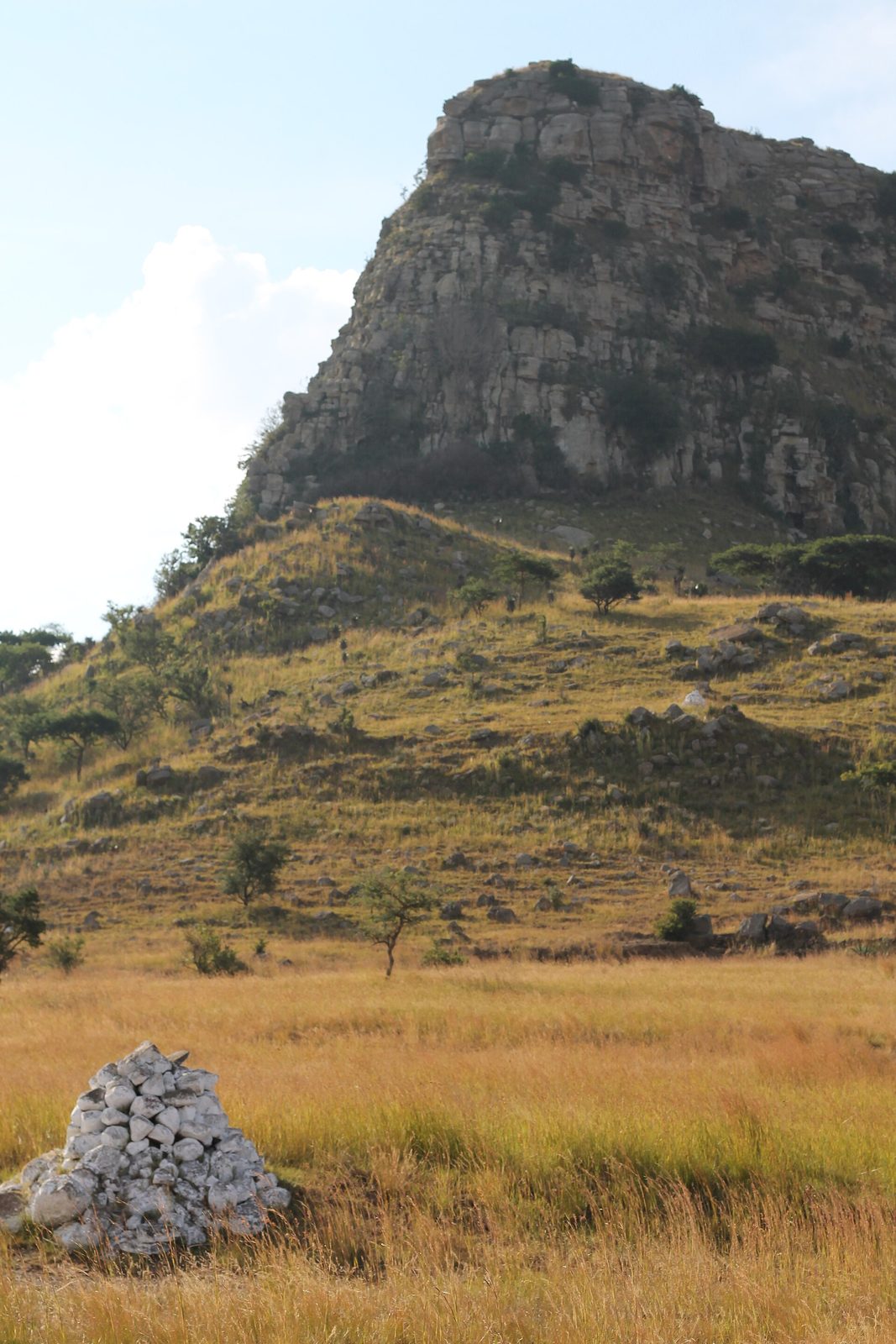 A white-painted stone cairn in dry grass, a steep rocky hill behind it.
