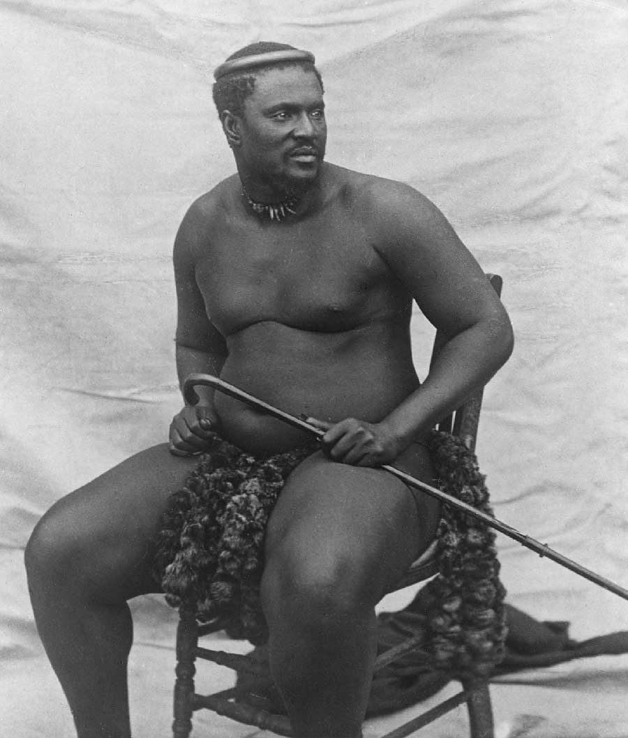 A black-and-white photograph of a heavy man seated on a chair, wearing a kilt of fur and holding a curved stick.